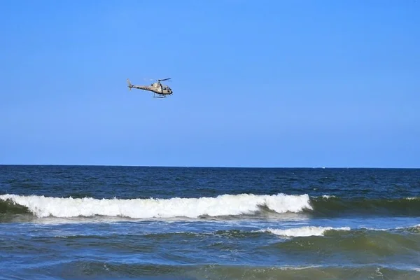 Foto colorida, helicóptero sobrevoando o mar.