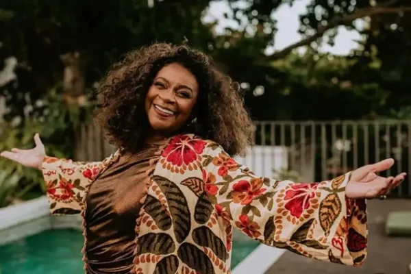 Imagem colorida mostra a cantora e atriz Zezé Motta. Ela tem os braços abertos e veste uma roupa com imagens de flores e folhas. Ela é uma mulher negra de cabelos cacheados e sorri - metrópoles