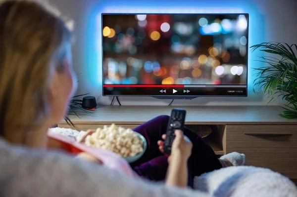 Mulher sentada num sofá com controle na mão assistindo TV