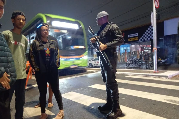 Imagens coloridas mostram um policial militar no centro, com um fuzil, parado sobre a faixa de pedestres, um ônibus próximo e moradores do lado esquerdo do PM, em protesto na via