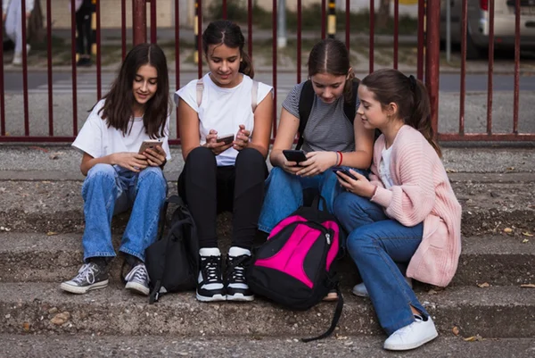 Foto colorida de quatro meninas sentadas em escada de escola usando celular - Metrópoles