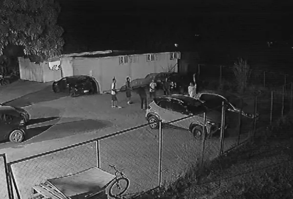 Imagem em preto e branco de jovens brincando no estacionamento de uma escola, com carros ao redor