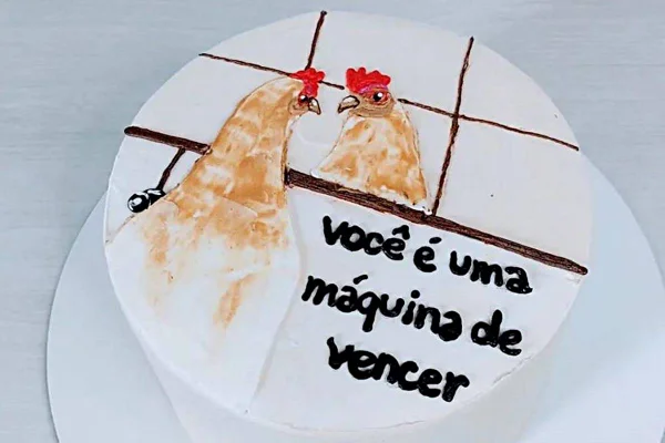 Na foto temos um bolo redondo branco com um desenho de uma galinha de cotar e outro dela de frente com frase você é uma maquina de vencer