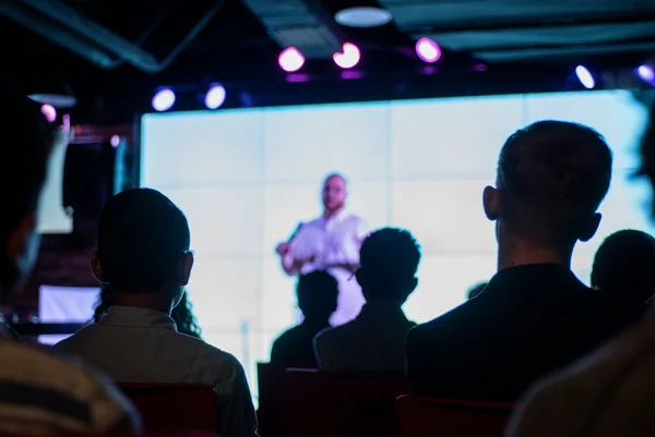 Fotografia colorida mostrando auditório cheio assistindo palestrante em palco com telão atrás - Metrópoles