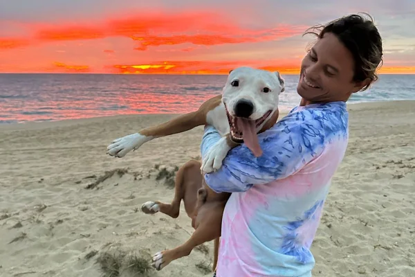 Imagem colorida de homem sorrindo e segurando cachorro de grande porte no colo - Metrópoles