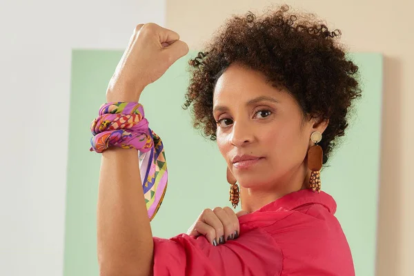 Campanha de Outubro Rosa da marca Cantão. Na foto é possível ver Jociane Monteiro, 45, criadora do projeto social Amigas do Peito Por Aí, e em remissão de tratamento contra o câncer de mama. Ela é uma mulher de meia idade negra com cabelo cacheado curto e usa uma camisa rosa de botão, além de um lenço estampado, também rosa, amarrado no pulso.