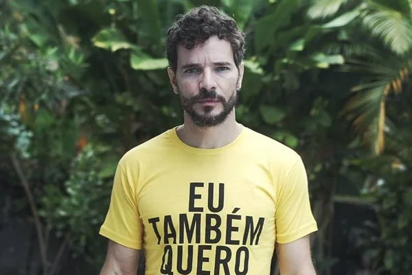 Foto colorida de Daniel de Oliveira com uma camisa amarela com os dizeres "eu também quero" e folhas verdes ao fundo - Metrópoles