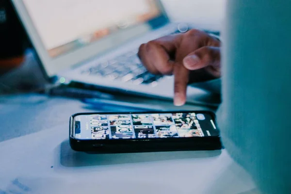 Pessoa mexendo no celular sob a mesa, ao lado de um laptop - Metrópoles