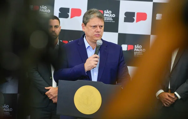 Imagem colorida mostra Tarcísio de Freitas, homem branco, grisalho, de terno azul escuro e camisa azul clara, falado ao microfone em um púlpito no palácio dos bandeirantes, entre câmeras - Metrópoles