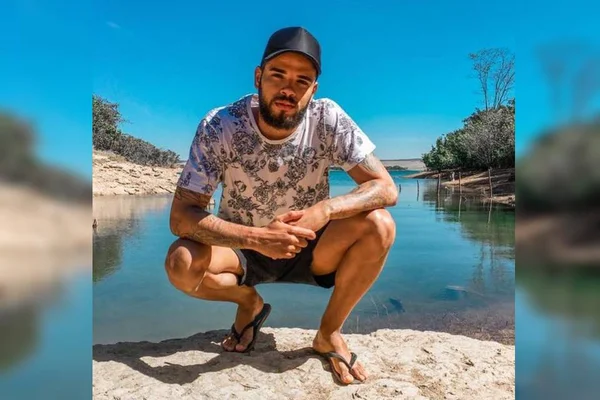 Alef Vogado. Jovem agachado a beira de rio com bermuda, camiseta estampada e boné preto - Metrópoles