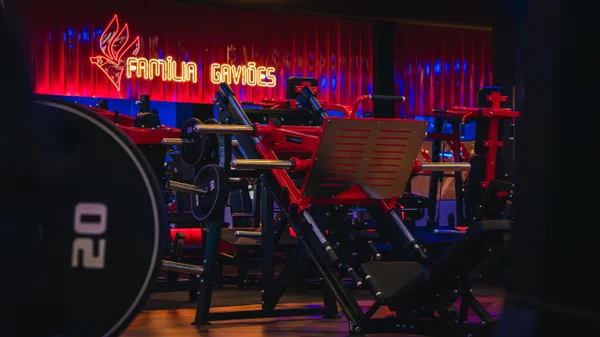 Área de musculação da academia, com equipamentos e pesos organizados no espaço. A iluminação em tons escuros e vermelhos reforça a identidade visual da unidade.
