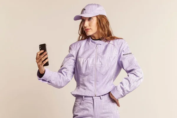 Mulher com roupas lilás, ela está com boné e tirando uma selfie com o celular - Metrópoles
