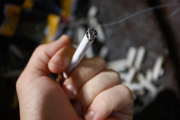 Mão segurando cigarro acesso, soltando fumaça. Ao fundo, pontas de cigarro e carteiras usadas jogadas no chão - Metrópoles