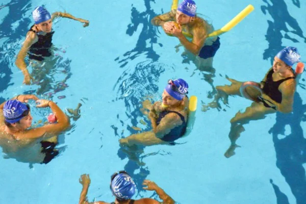 Imagem colorida feita do alto mostra seis pessoas dentro de uma piscina. Elas estão boiando sentadas com a ajuda de boias e conversam entre si. Elas vestem maiô ou sunga e toucas na cabeça - Metrópoles