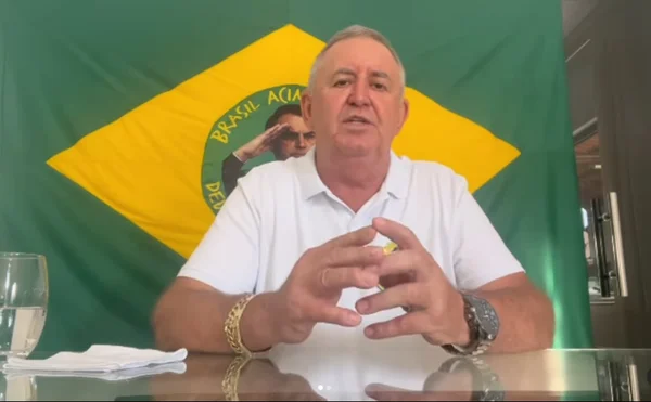 Empresário acusado de assédio eleitoral na Bahia aparece dizendo que se tratava de uma "brincadeira". Na foto ele está numa mesa, falando, com uma bandeira do Brasil personalizada com o presidente Bolsonaro atrás - Metrópoles