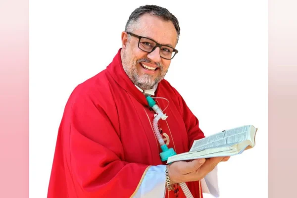 Imagem de fundo borrado mostra padre com vestimenta vermelha e com a bíblia na mão. Ele tem traqueostomia por sua doença rara - Metrópoles