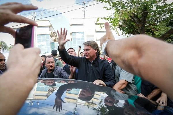 Fotografia colorida mostra homem com blusa de frio preta apoiado em carro com mão esquerda e com a mão direita estendida para pessoas que o cercam