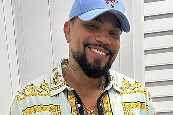 Naldo posa, sorridente, de camisa com estampa amarela e boné azul - Metrópoles