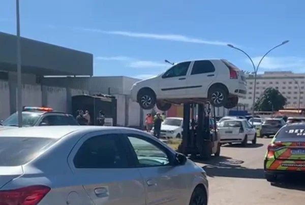 Imagem de um carro branco sendo erguido por uma empilhadeira em meio a um estacionamento