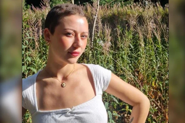 Foto colorida de mulher jovem com cabelos raspados e blusa branca em cenário parecido com um campo, plantas e mato - Metrópoles.