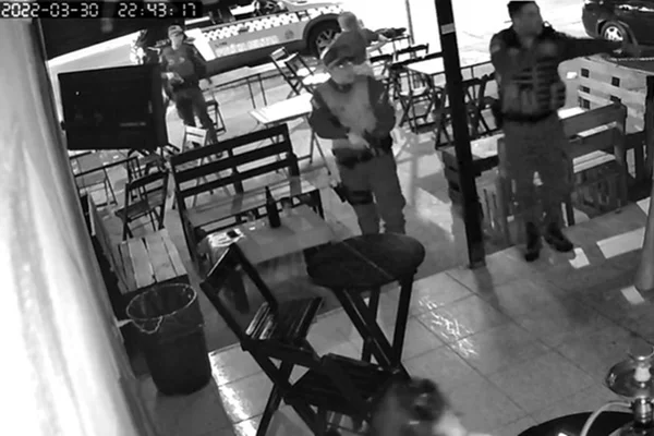 Imagem em preto e branco de policiais armados em bar com mesas e cadeiras