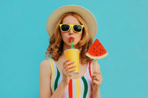 Mulher de oculos, chapéu com melancia na mão e bebendo suco