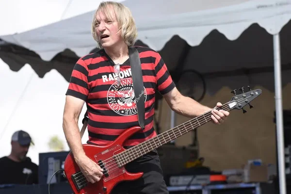 Rick Anderson. Ele segura uma guitarra vermehla, está com um ablusa listrada em vermelho e preto, tem pele clara e cabelos loiros na altura dos ombros - Metrópoles