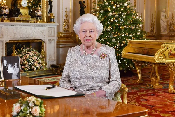 Mulher com cabelos brancos sentada. Ao fundo, uma árvore de Natal e um piano. Na mesa, há um porta-retrato