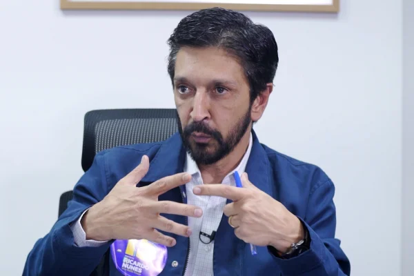Imagem colorida mostra Ricardo Nunes, homem branco, de cabelo e barba pretos, vestindo jaqueta azul e camisa branca, sentado em uma cadeira com uma parede branca ao fundo, conversando - Metrópoles