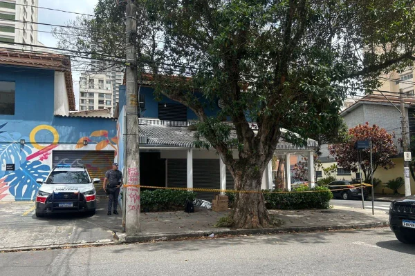 Imagem colorida mostra área de calçada isolada com fita colorida em laranja e vermelho. Um carro de polícia está estacionado e um policial fardado está ao lado - Metrópoles