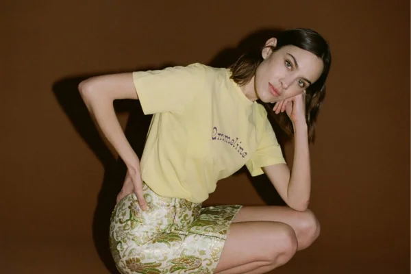 A modelo Alexa Chung usa camiseta amarela e saia com estampa de cobra. Ela está agachada, posando para foto, em um fundo cor de vinho.