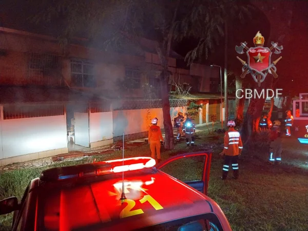 Foto colorida tirada à noite de casa iluminada por luz externa vermelha e homem com uniforme de bombeiro o