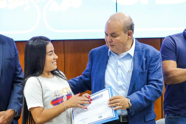 Foto colorida de homem careca de terno entregando escritura a jovem de cabelos pretos e roupa casual