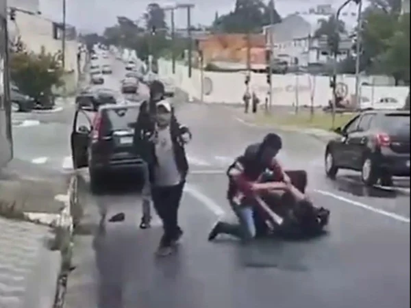 Imagem colorida mostra dois adolescentes brigando amontoados no chão enquanto um homem segura uma arma
