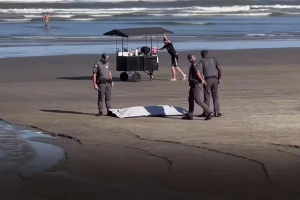 Foto colorida de corpo de adolescente morta estendido na areia da Praia Grande, em frente ao mar, com policiais militares ao redor - Metrópoles