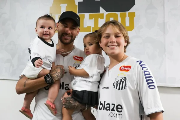 Foto colorida de homem segurando duas crianças no colo. Tem um menino ao lado deles - Metrópoles