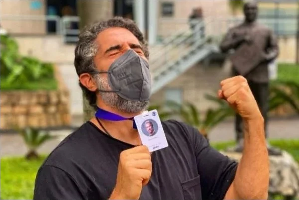 Na imagem colorida, um homem está posicionado no centro. Ele usa máscara e camiseta preta
