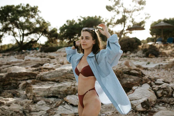 Mulher branca e jovem, de cabelo castanho liso, posando para foto perto de pedras em um praia. Ela usa um top e uma calcinh de biquíni, ambos marrons, e uma camisa de botão azul. - Metrópoles