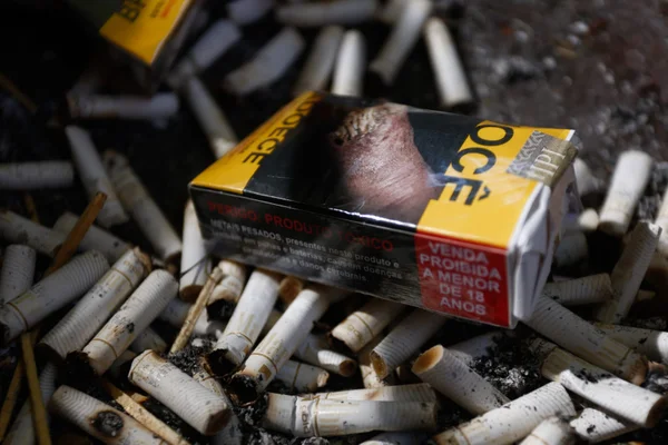 Foto de carteira de cigarro amassada jogada sob vários cigarros usados e queimados, com cinzas por cima - Metrópoles
