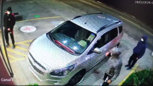 imagem colorida mostra cena filmada por câmera de segurança. três homens com moletons e máscaras estão ao redor de um carro de cor clara no estacionamento de uma farmácia - metrópoles