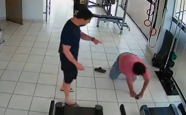 Homem de preto em pé apontando o dedo para homem de camisa rosa abaixado no chão em academia