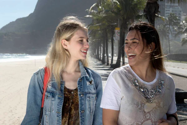 Duas mulheres, uma loira e uma morena, caminhando aos risos na praia - Metrópoles