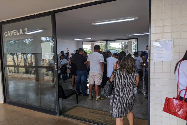 pessoas dentro de uma sala de vidro reunidas em uma reunião que aparenta ser um velório