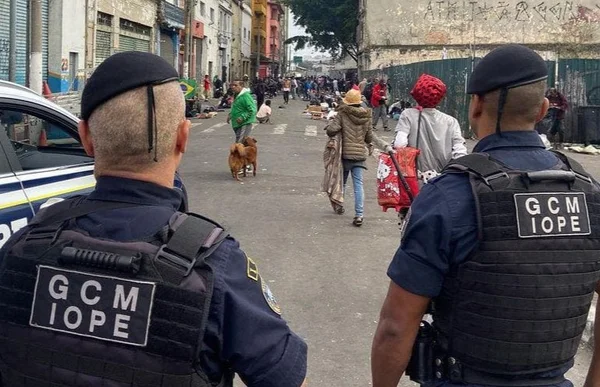 Imagem colorida mostra dois agentes da CGM/IOPE de costas; ao fundo, usuários de drogas transitam em rua estreita - Metrópoles