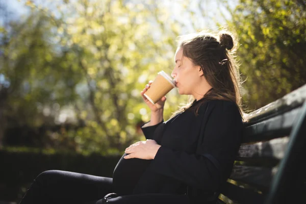 Imagem mostra gestante com a mão na barriga, tomando uma bebida em um copo para viagem e sentada em um banco - Metrópoles
