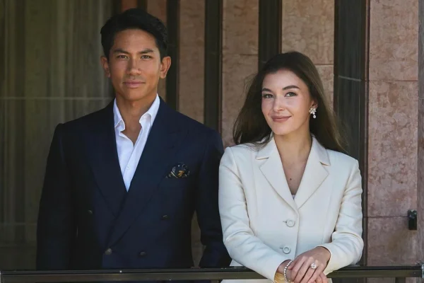 Foto de homem, vestido com blazer azul e blusa branca, ao lado de mulher com blazer nude. Ela está mostrando um anel. Os dois estão em uma varanda - Metrópoles