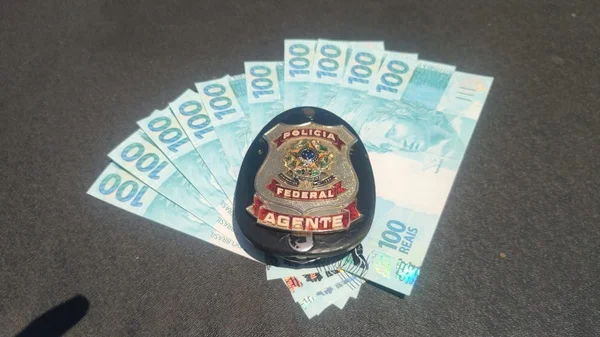 Imagem colorida mostra dez notas de R$ 100 na frente de um distintivo da Polícia Federal; as notas falsas foram descobertas dentro de uma correspondência direcionada a um adolescente - Metrópoles