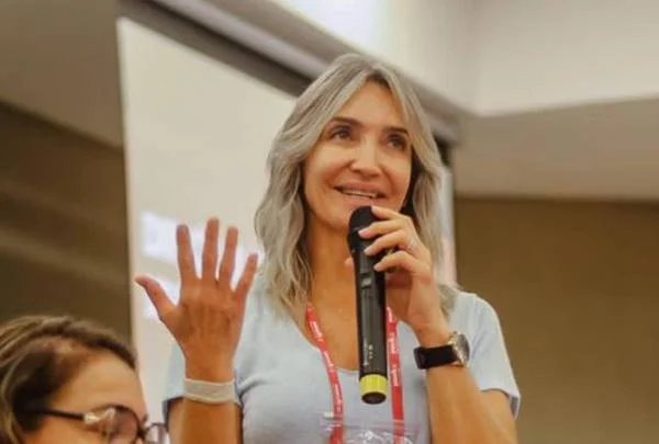 Fotografia colorida de mulher com camiseta cinza segurando microfone com a mão esquerda e mostrando mão direita