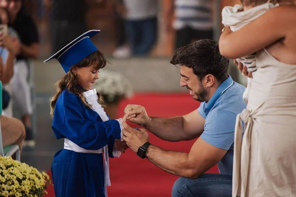 Foto colorida de um pai entregando um anel de formatura para a filha pequena - Metrópoles