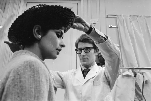 Na imagem, de 1965, em preto e branco, o estilista Yves Saint Laurent ajusta um chapéu na cabeça de uma modelo - Metrópoles
